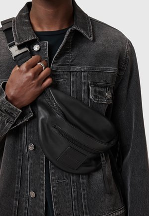 Borsa a tracolla in pelle nera con tasca frontale con zip, forma curva e patch con logo. Indossata sopra una giacca di jeans grigia.