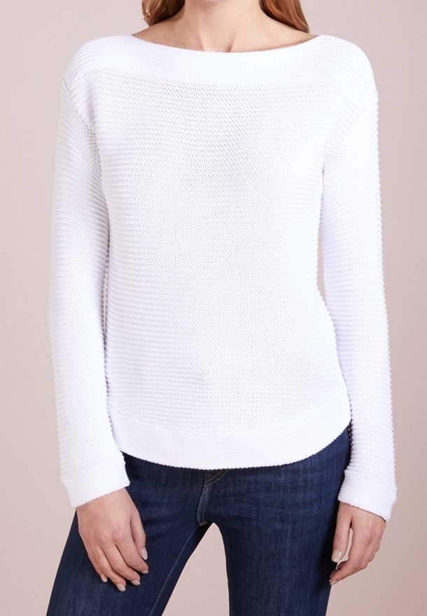 Femme portant un pull en tricot texturé blanc avec un col bateau et des manches longues, associé à un jean bleu foncé, se tenant devant un fond neutre.