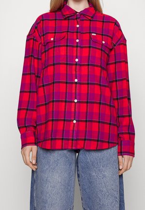 Persoon draagt een rood-paars geruit overhemd met knopen en twee borstzakken, gecombineerd met blauwe wijde jeans.