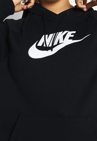 Person som justerar huvan på en svart hoodie med en stor vit Nike-logotyp och swoosh på bröstet.