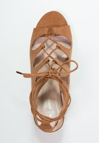 Hnědé semišové sandály s otevřenou špičkou, zkříženými popruhy a ozdobnou mašlí. Mají plochou podrážku a hladký vnitřní povrch.