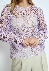 Top in pizzo lavanda con motivi intagliati intricati e maniche lunghe, indossato sopra un indumento beige neutro. Tessuto morbido e texturizzato.