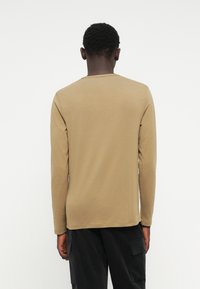 Camicia a maniche lunghe in tessuto beige, con scollatura a girocollo, texture liscia e design aderente. Cuciture visibili lungo l'orlo e le maniche.