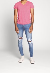 Röd v-ringad t-shirt i kombination med slitna blå skinny jeans och vita sneakers. Tillverkad av bomull, avslappnad passform, märkbara hål i jeansen.