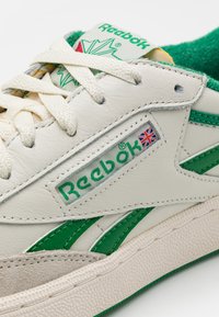 Beli usnjeni športni copat z zelenimi detajli in teksturiranim gumijastim podplatom. Ima jezik v zeleni barvi in logotip Reebok z detajlom britanske zastave.