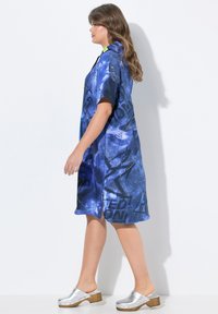 Abito camicia blu con grafica e maniche corte, caratterizzato da una combinazione di stampe e accenti gialli a contrasto, abbinato a zoccoli argentati su base di legno.
