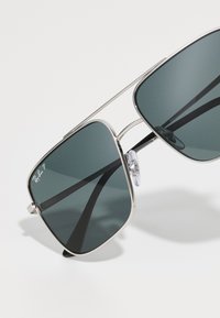 Aurinkolasit tummanvihreillä linsseillä, metallinhohtoisella hopeakehyksellä, suorakulmaista muotoa ja säädettävät nenätyynyt. Linsseissä on Ray-Ban P -logo.