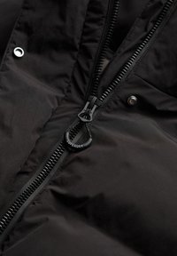La chaqueta aislante negra cuenta con un tejido liso y duradero, un prominente zipper negro y un tirador circular con la marca, con una superficie texturizada.