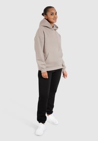 Hoodie beige surdimensionné en tissu doux avec une poche frontale, associé à un pantalon noir et des baskets blanches. Texture lisse, coupe décontractée.