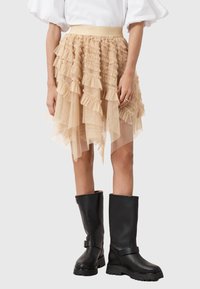 Gonna in tulle beige a strati con dettagli arricciati, caratterizzata da un orlo asimmetrico. Indossata con stivali in pelle nera fino al ginocchio.