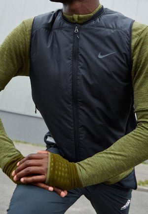 Henkilö, joka pukee mustan, hihattoman Nike-liivin vihreän pitkähihainen paita, jossa on peukaloaukot, venyttelee ulkona päällystetyllä alueella.