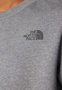 Grijze sweatshirt met een zachte textuur en geribbelde halslijn, met een zwart logo van "The North Face" op de linkerkant van de borst.