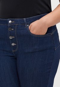 Jeans de mezclilla azul oscuro con cintura alta, cremallera de cinco botones y bolsillo lateral. Presentan costuras visibles y una textura suave.