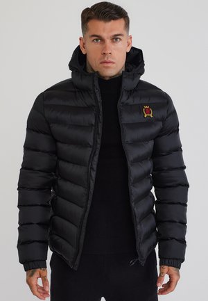 Uomo con una giacca imbottita nera con cappuccio, in piedi e che guarda dritto davanti a uno sfondo uniforme.