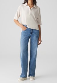 Beige pikétröja med korta ärmar och knappslå, i kombination med ljusblå raka jeans och vita skor. Enkelt mönster och textur.