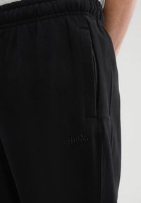 Czarne dresy wykonane z miękkiego materiału, z elastycznym paskiem w talii, bocznymi kieszeniami oraz subtelnym wytłaczanym logo na dolnej części nogawki.