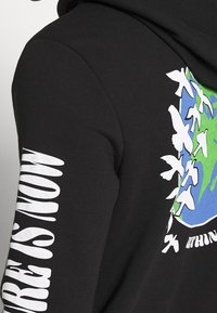 Sudadera negra con un gráfico de un globo terráqueo en verde y azul y siluetas de pájaros en blanco. Presenta el texto "LA NATURALEZA ES AHORA" en la manga. Textura suave.
