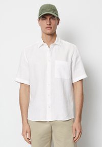 Homme portant une chemise blanche à manches courtes avec des boutons, un pantalon beige et une casquette de baseball olive, regardant vers l'avant.