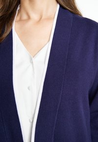 Cardigan navy con rifiniture bianche, texture morbida, design aperto e blusa bianca a V con dettagli dei bottoni sotto.