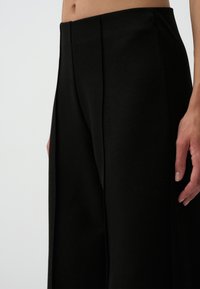 Pantalon noir ajusté avec une taille haute, des plis latéraux et une texture lisse. Présente une coupe droite, offrant une silhouette élégante.