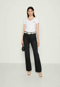 Blusa curta de mangas com um pequeno logótipo, jeans flare de cintura alta em denim escuro, cinto branco e mala preta. O modelo usa sandálias brancas de salto.