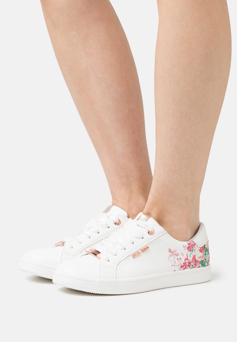 Paire de baskets basses blanches avec imprimé floral sur les côtés et détails en or rose, portées sur des jambes nues sur un fond uni.