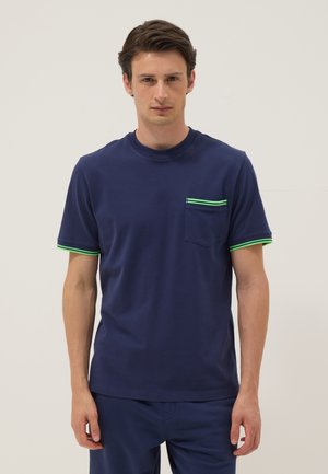 Man wearing navy blue T-shirt with neon green trim on pocket and sleeves, standing against plain light background.