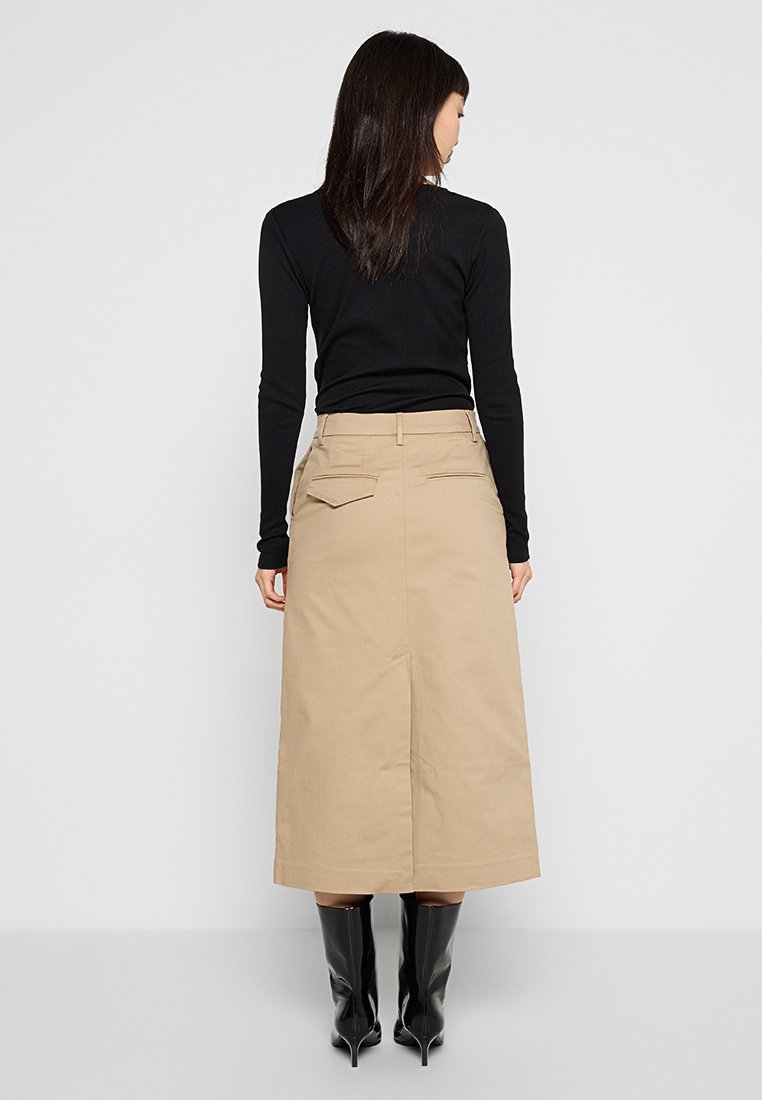 Beżowa spódnica do kolan z centralnym rozcięciem z tyłu, dwoma tylnymi kieszeniami i usztywnionym paskiem w talii, w połączeniu z czarną bluzką z długim rękawem i wysokimi butami na obcasie.