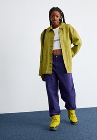 Zelená oversized bunda, bílý crop top, fialové volné kalhoty, žluté texturované boty s dekorativními šňůrkami. Jednoduché pozadí, vnitřní prostředí.