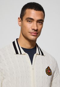 Cardigan de punto en crema con un patrón de trenzas, cuello a rayas en blanco y negro, cremallera completa y emblema bordado en el pecho.