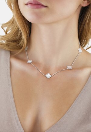 Frau trägt eine silberne Halskette mit fünf kleeblattförmigen weißen Steinanhängern, beiges Oberteil, sichtbarer Hals und oberer Brustbereich.