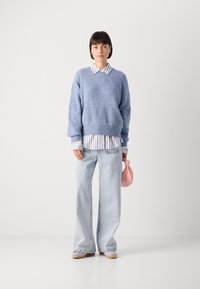 Blue knitted sweater over a white and blue striped shirt, paired with light blue wide-leg jeans and holding a small pink handbag.