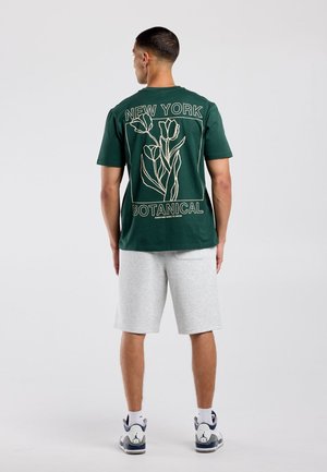 Man staat met zijn rug naar de kijker, draagt een donkergroen T-shirt met bloemenprint en tekst "New York Botanical" en lichtgrijze korte broek en sneakers.