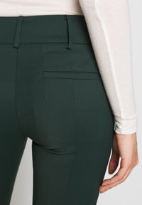 Mörkgröna skräddarsydda byxor i ett mjukt, stretchigt material. Har en bakficka och bälteshällor, tillsammans med en ljusfärgad ribbad topp.