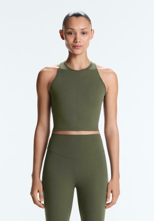 Kvinde står og vender fremad iført en olivengrøn ærmeløs crop-top og matchende højtaljede leggings mod en ensartet lys baggrund.