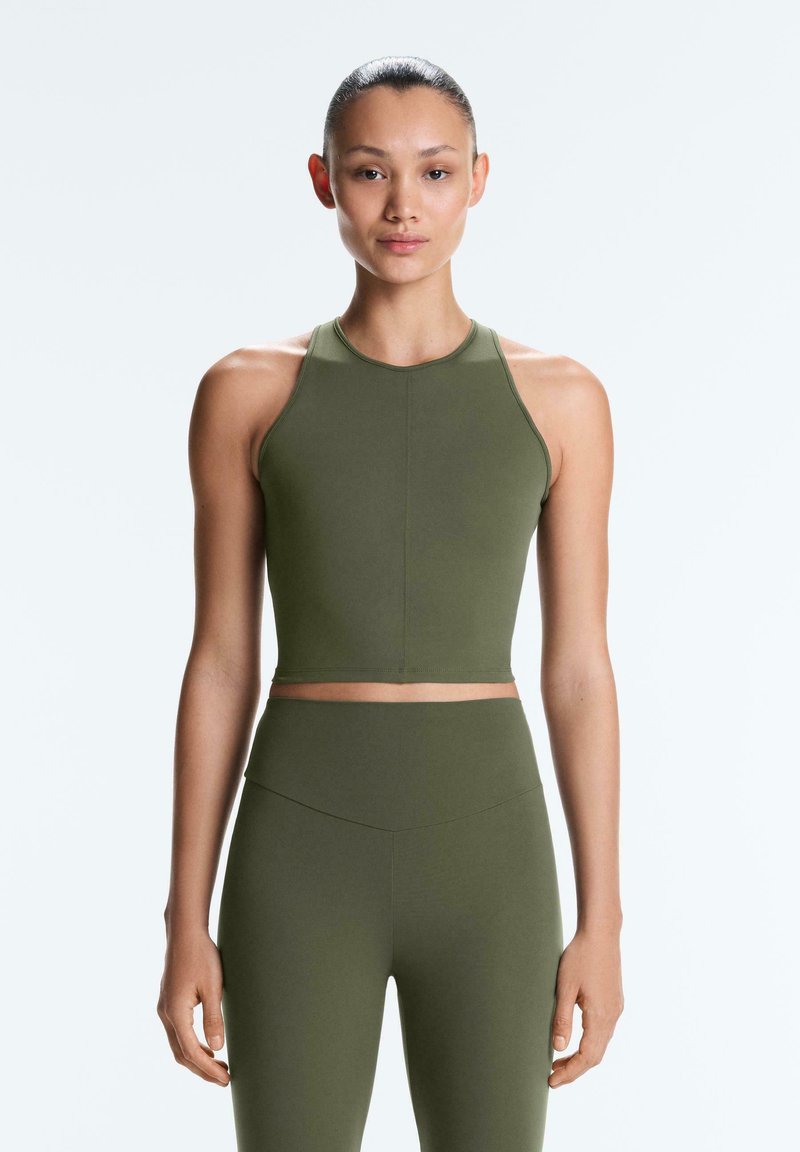 Frau steht mit dem Gesicht nach vorne, trägt ein ärmelloses, olivgrünes Crop-Top und passende, hoch taillierte Leggings vor einem schlichten hellen Hintergrund.