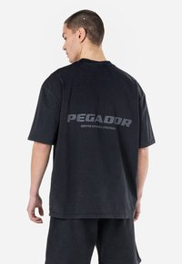 Juodas perdidėlis t-shirt su trumpomis rankovėmis, su pilku atspaudu „PEGADOR“ ant nugaros, pagamintas iš minkštos medvilnės.