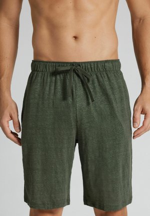 Midterparti af person iført mørkegrønne elastiske shorts med snøre, stående mod en ensfarvet grå baggrund.