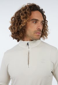Vyras su garbanotais plaukais, vilkintis kreminės spalvos zip-up švarką su užrašu "PROTEST" ant užtrauktuko, žiūrintis į šoną prieš baltą foną.