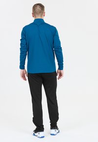 Herrträningsklädsel bestående av en långärmad teal-färgad topp och svarta byxor. Toppen har en jämn textur, medan byxorna är lätta och utformade för att smalna av.