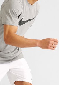 Grå kortärmad idrottströja med svart Nike Swoosh-logga, i kombination med vita shorts. Tygmaterialet verkar lätt och strukturerat.