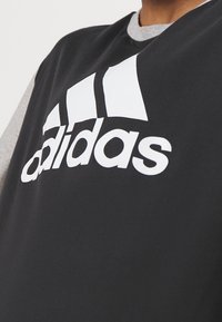 Černé atletické tričko s bílým logem "adidas". Má šedý kulatý výstřih a šedé rukávy. Látka vypadá hladce s mírným leskem.