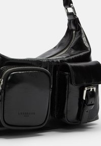 Czarna skórzana torba o błyszczącej fakturze, z kieszenią na zamek błyskawiczny i akcentem w postaci klamry. Owalny kształt z wieloma kieszeniami.