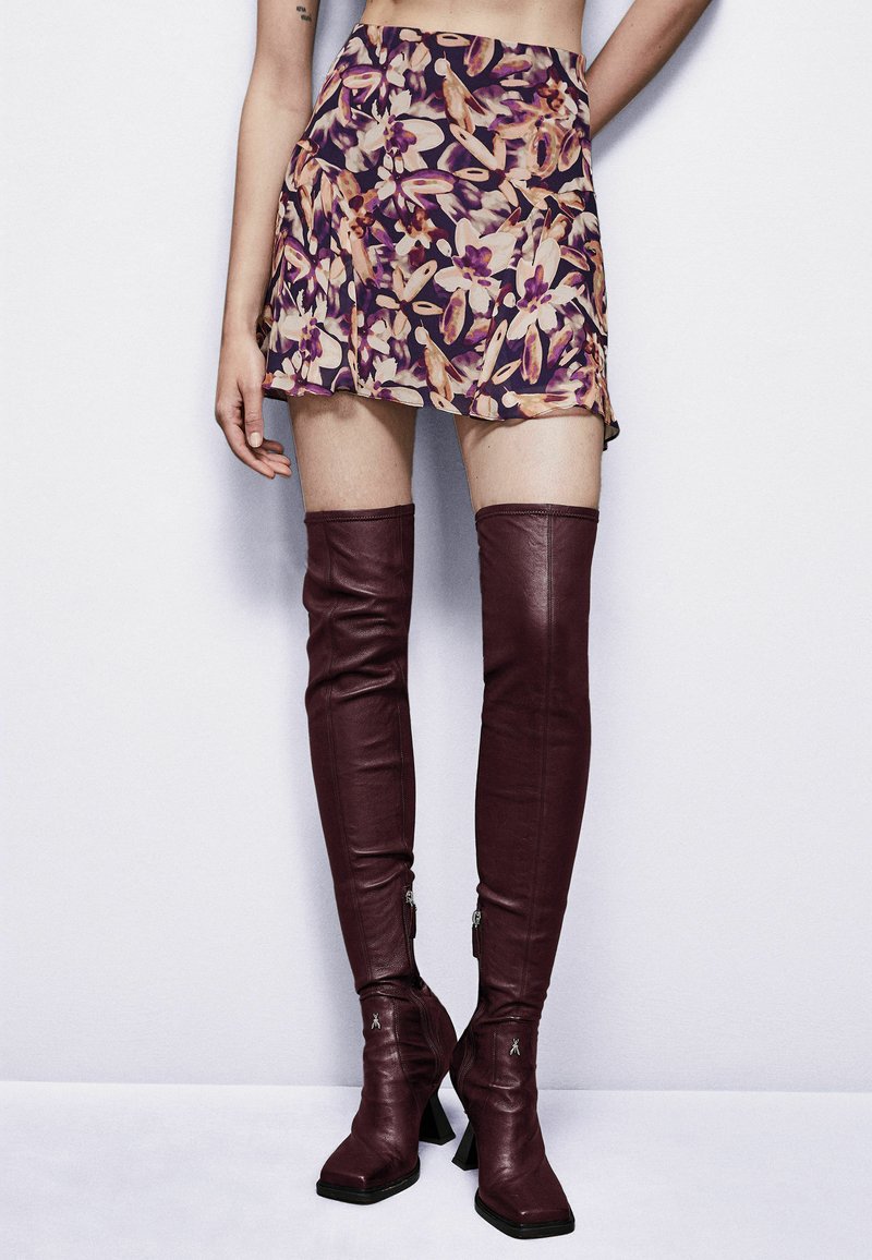 Stivali alti in pelle marrone con tacco a blocco e chiusura con zip, abbinati a una minigonna con motivo floreale nei toni del viola, rosa e crema.