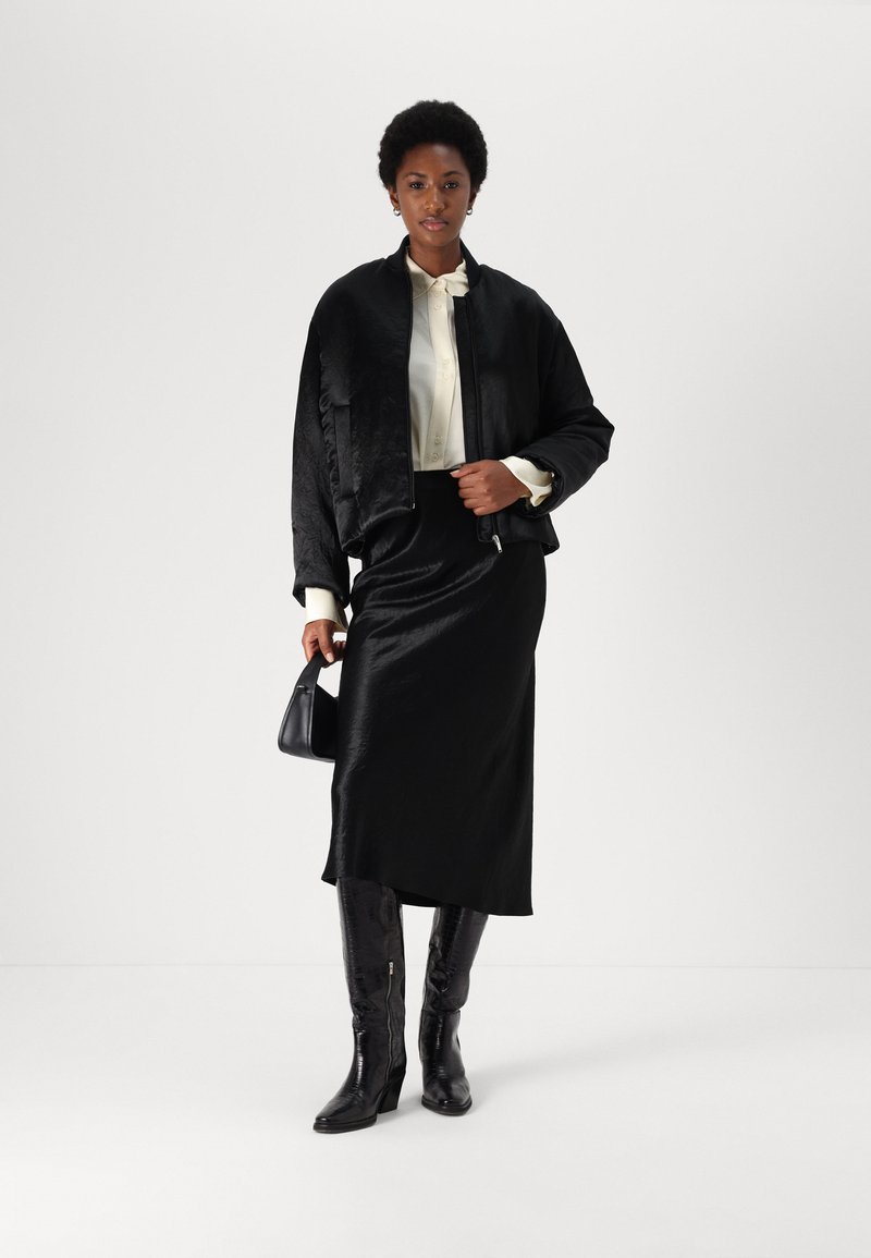 Veste bombardier en satin noir sur un chemisier crème, associée à une jupe en satin noir et des bottes noires texturées hauteur genoux. Tient un petit sac noir.