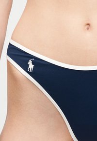 Κοντινό πλάνο της μέσης ενός ατόμου που φοράει μαγιό μπικίνι σε ναυτικό μπλε με λευκή επένδυση και μικρό λευκό λογότυπο παίκτη πόλο στη αριστερή πλευρά.