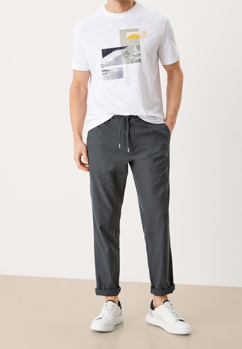 Homme portant un t-shirt blanc à motif graphique, un pantalon gris foncé avec cordon et ourlets, et des baskets blanches avec des détails noirs, debout les mains dans les poches.