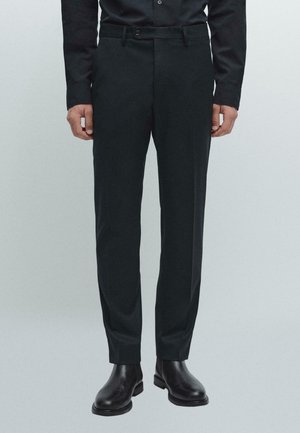 Pantalons taillés gris foncé avec une texture lisse, poches avant et fermeture à un bouton. Associés à des bottines en cuir noires.
