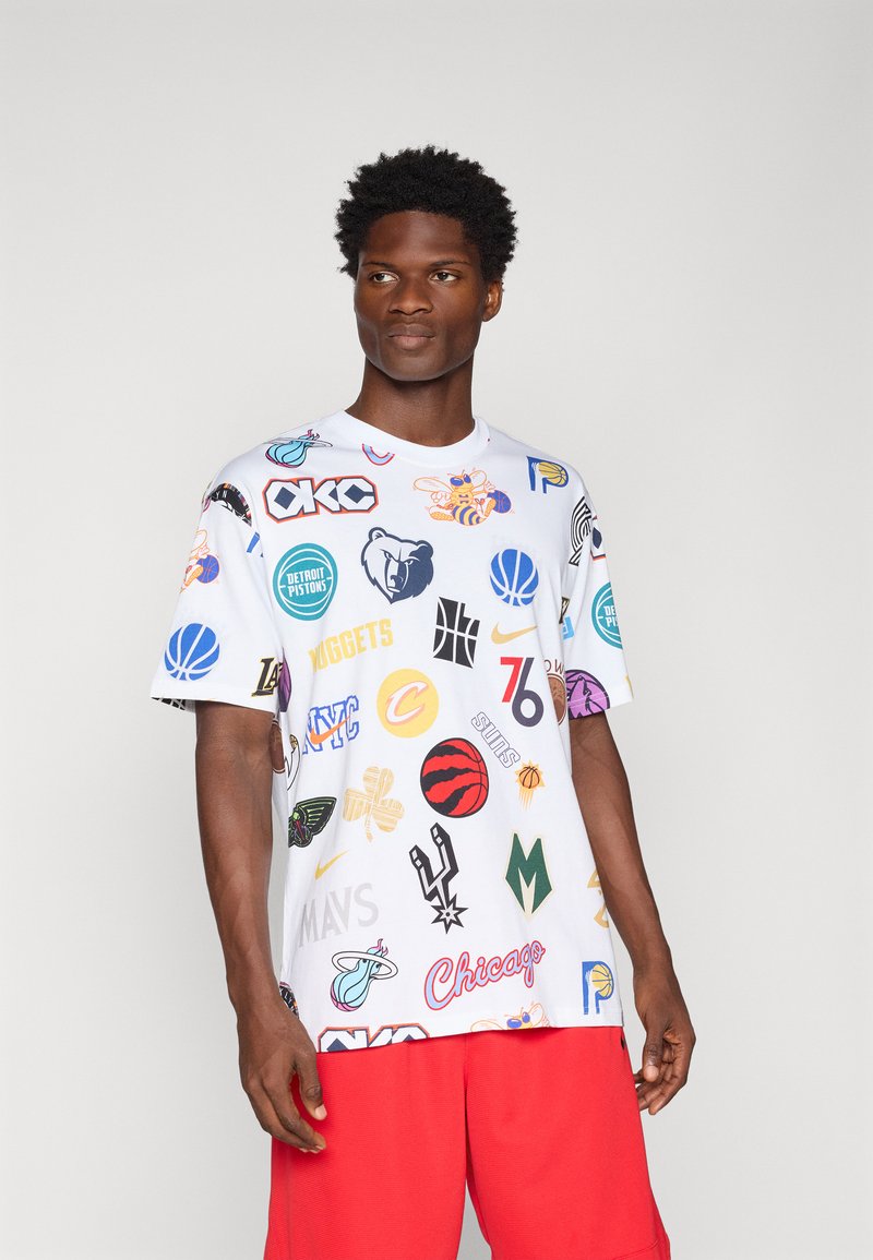 Homme portant un T-shirt blanc couvert de logos colorés d'équipes NBA et un short rouge, debout devant un fond clair uni.