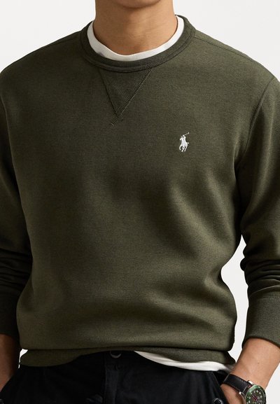 Olivengrønn genser med rund hals, med en hvit brodert logo på venstre bryst og ribbede mansjetter. Lagd over en hvit skjorte.
